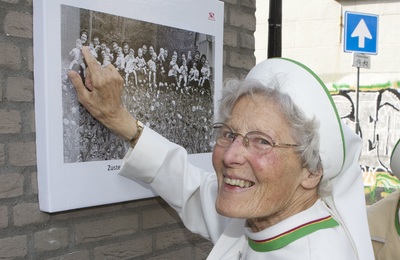 878102 Portret van een Zuster Augustines wijzend op het zojuist aangebrachte fotobord ter gelegenheid van het 80-jarig ...